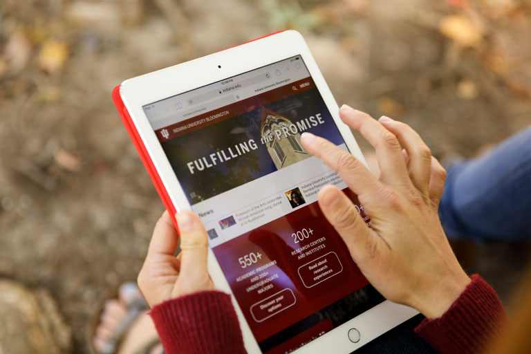 An IU Studios staff member presents a website on a tablet; the page shows the tower of the Student Building on the IU Bloomington campus along with the words 'Fulfilling the Promise' and some statistics