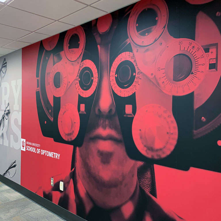 An Indiana University School of Optometry wall graphic depicts a person seated at a phoropter, an instrument used in eye exams
