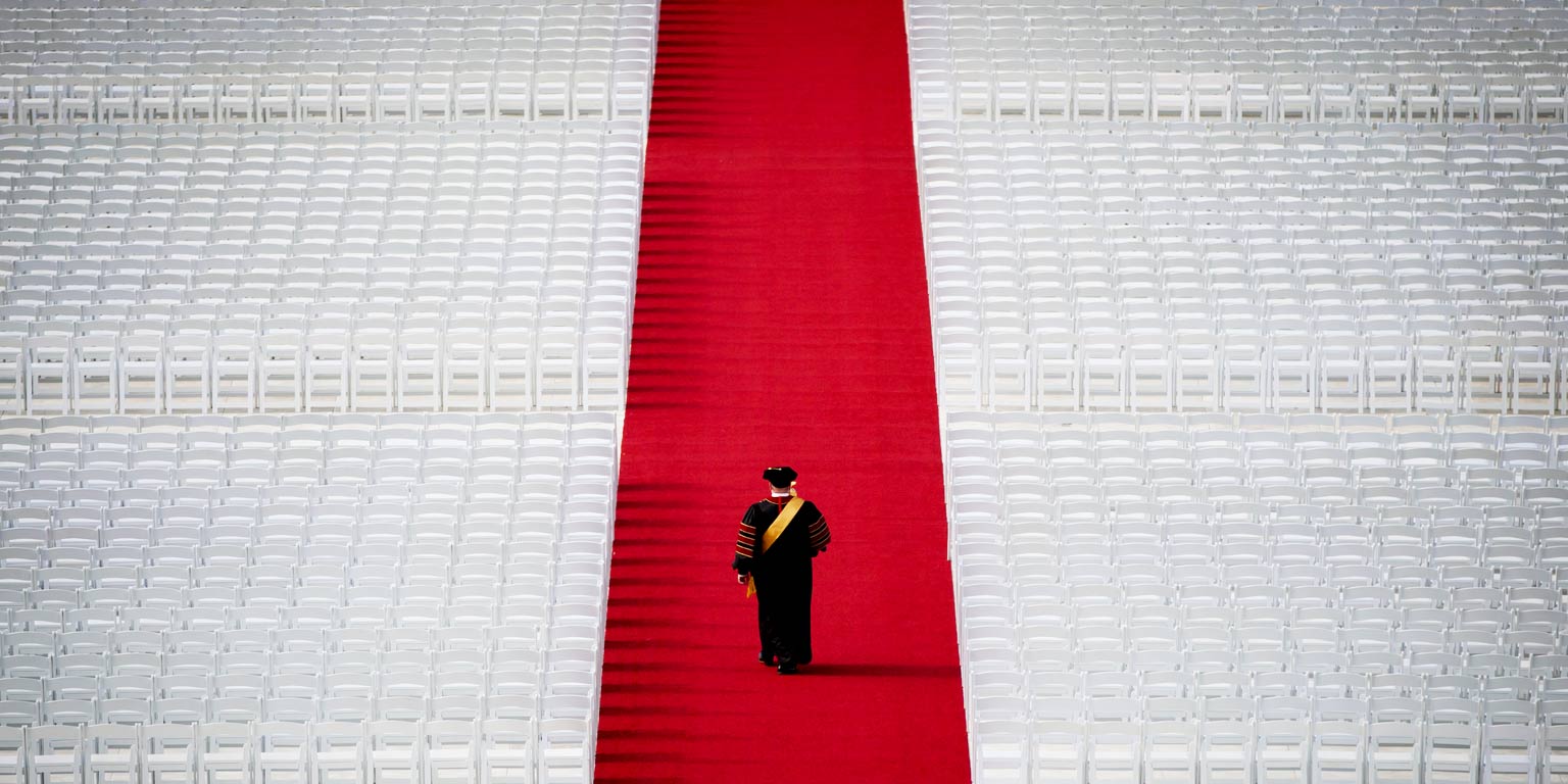 An administrator dressed in graduation regalia walks the red carpet in an empty stadium
